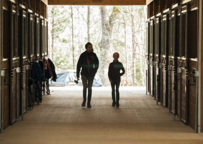 Horse stable entrance with two people walking inside, surrounded by wooden stalls and outdoor forest.
